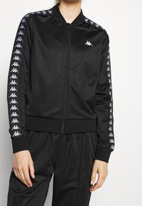 Svart zip-up jacka med ribbade ärmslut och krage, prydd med ett upprepat logomönster i ljuslila längs båda ärmarna. Slät, lättviktig tyg.