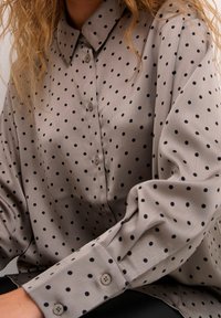 Blusa gris con lunares negros, de manga larga y con cierre de botones en el frente. Está hecha de una tela suave, con un cuello clásico y puños de botón.