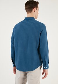 Cămașă cu mâneci lungi de culoare bleu închis, dintr-un material texturat. Prezintă un guler clasic, manșete cu nasturi și un tiv drept cu dungi verticale subtile.