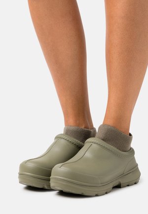 Groene rubberen klompen met een gestructureerde bovenkant, gestikte afwerking en een zachte, gebreide kraag. Chunky buitenzool voor grip en comfort.