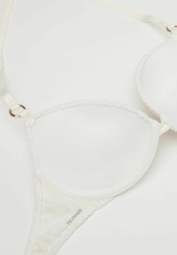 Sostén blanco acolchado con textura suave, bordes de encaje, tirantes ajustables y acentos en oro. Presenta un branding sutil.