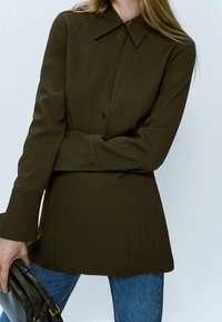 Chemise à manches longues vert foncé avec un col structuré, fermeture à boutons sur le devant et détails plissés. Portée avec un jean bleu et un sac noir.