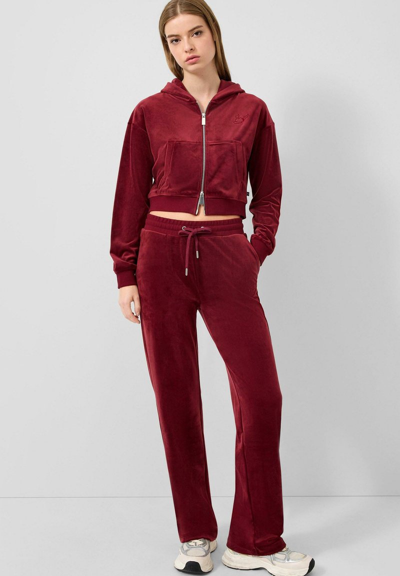 Jongeman in een bordeaux velours hoodie met rits en bijpassende joggingbroek met een trekkoord, met witte sneakers, die tegen een effen lichtgrijze achtergrond staat.