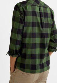 Persona che indossa una camicia a maniche lunghe a quadretti verdi e neri con le maniche arrotolate e pantaloni beige, vista da dietro.