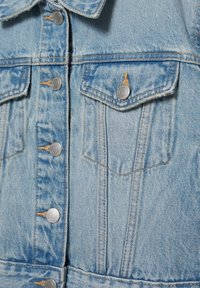 Veste en jean bleu clair avec boutons-pression en métal et poches sur la poitrine, mettant en valeur les coutures détaillées et la texture.