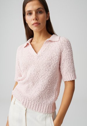 Femme portant un pull à manches courtes texturé rose clair avec col et un pantalon blanc, debout devant un fond gris uni.