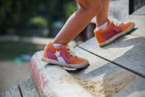 Child wearing orange and pink sneakers climbs a wooden inclined surface outdoors in a play area.
