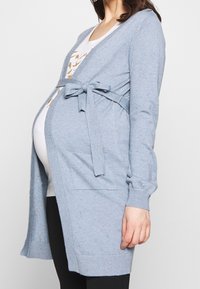 Modrý pletený cardigan má v pase zavazovací pásek, žebrované manžety a kapsy. Materiál má skvrnité texturování.