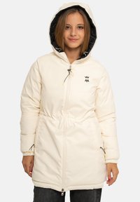 Jeune femme portant un manteau d'hiver à capuche de couleur crème avec une doublure noire, les mains dans les poches, debout devant un fond uni.