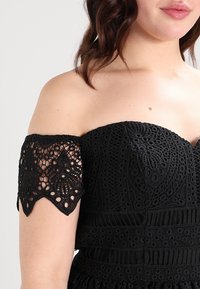 Černé šaty bez ramínek s propracovanými krajkovými rukávy ve tvaru vln, texturovaným tělem s geometrickými vzory a přiléhavým pasem.