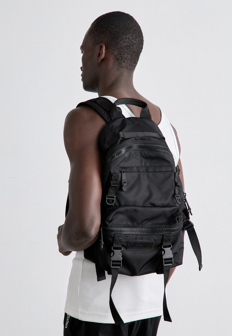 Svart nylonryggsäck med flera zippered fack, justerbara axelremmar och logodetalj på framsidan. Texturerad yta kontrasterar med slät tyg.