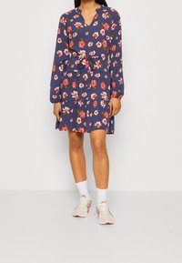 Blommig klänning med marinblå bakgrund, långa ärmar, V-ringning och knytmidja. Har en flerlagrad kjol och röda blommönster. Bärs med sneakers.