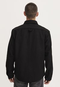 Camisa negra de botonadura con cuello puntiagudo, mangas largas y corte recto. Presenta un lazo en la parte posterior y puños abotonados.