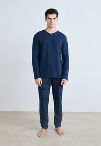 Camisa de manga larga azul marino con cuello en v combinada con pantalones de pijama azul marino de diseño estampado, que presentan un patrón de diamantes. El modelo está descalzo sobre un suelo claro.