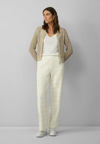 Beige gebreide cardigan, lichtgewicht, met patroon; witte mouwloze top; gestructureerde crèmekleurige wijde broek; witte sneakers.