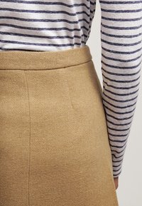 Jupe en tissu tissé brun avec une texture lisse, taille haute, et détail de couture sur le côté, associée à une chemise à manches longues rayée bleu et blanc.