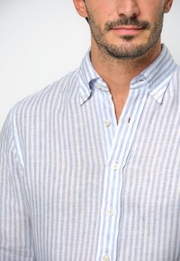 Lekka lniana koszula w niebiesko-białe pionowe paski, z kołnierzykiem zapinanym na guziki i białymi guzikami. Miękka faktura i swobodny krój.