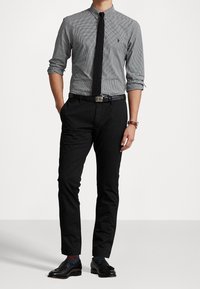 Muž v černých mokasínech, černých kalhotách, černém pásku, šedé vzorované košili s vyhrnutými rukávy, černé kravatě a hodinkách, stojící s rukama v kapsách.