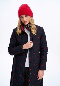Čierna prešívaná bunda s červenými bodkami, s vysokým golierom a predným zapínaním na cvoky. Nosená s červenou pletenou čiapkou.