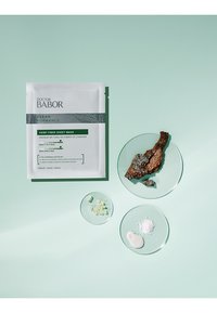 Hemp Fiber Sheet Mask package in white with green accents. Accompanied by glass plates containing green capsules, brown bark, powder, and cream.