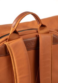 Zaino in pelle tan con una superficie liscia, dotato di manico superiore e spallacci imbottiti. Include un’etichetta con logo sul retro.