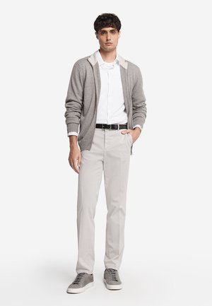 Mężczyzna ubrany w szarą bluzę z kapturem i zamkiem na białą koszulę, beżowe spodnie z czarnym paskiem i szare trampki, stoi z ręką w kieszeni.