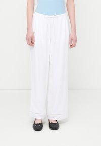 Γυναίκα που φοράει λευκό, φαρδύ παντελόνι με κορδόνι και μαύρα μπαλαρίνες, στέκεται μπροστά σε απλό λευκό φόντο.