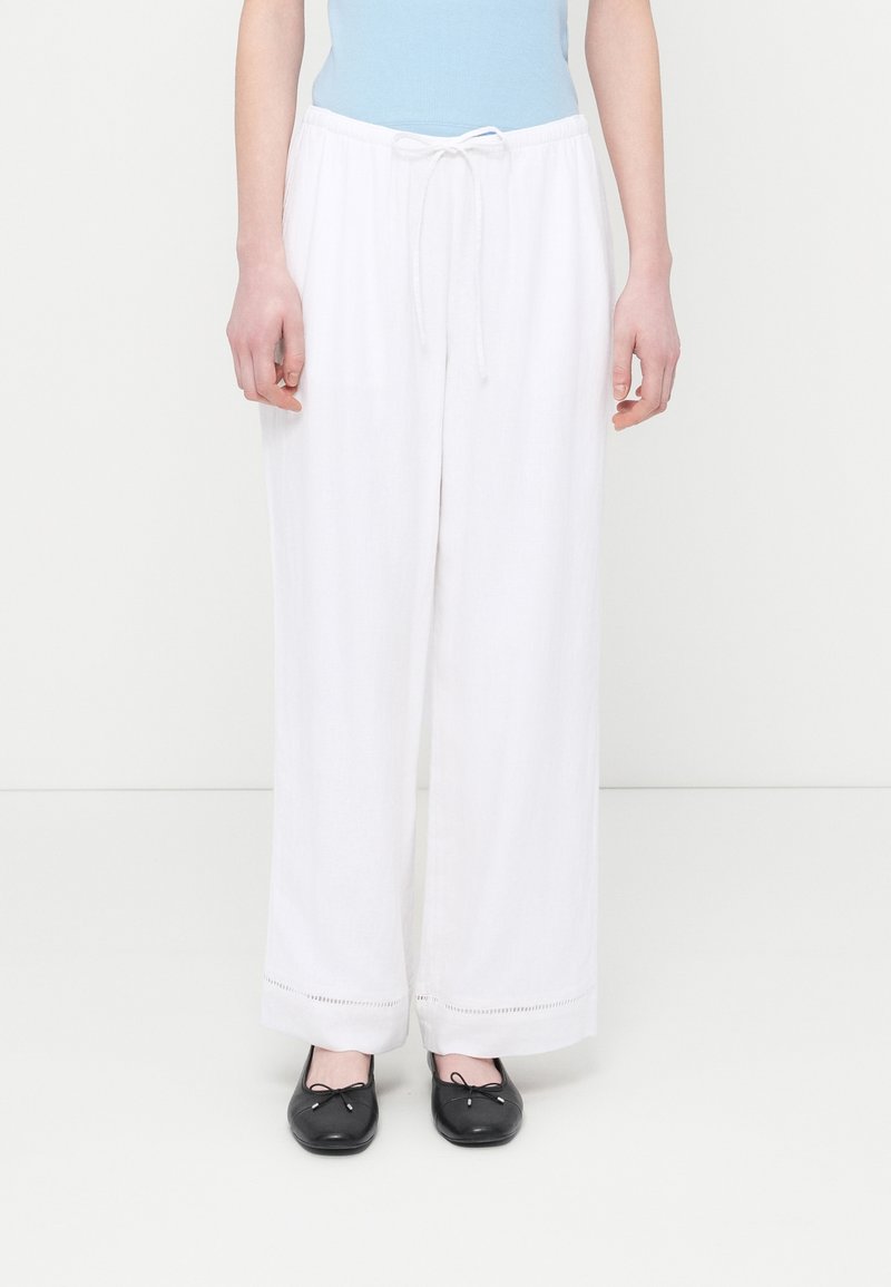 Γυναίκα που φοράει λευκό, φαρδύ παντελόνι με κορδόνι και μαύρα μπαλαρίνες, στέκεται μπροστά σε απλό λευκό φόντο.