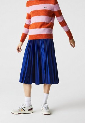 Personne portant un pull rayé orange et rose, une jupe midi plissée bleue, des chaussettes blanches et des baskets blanches sur un fond uni.