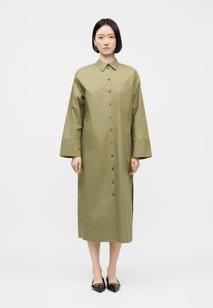 Kobieta z krótkimi czarnymi włosami, ubrana w długą oliwkowozieloną sukienkę koszulową na guziki oraz czarne spiczaste baleriny, stojąca na tle jednolitej białej ściany.