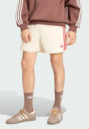 Mężczyzna w beżowych szortach z czerwonymi paskami, brązowym swetrze z białymi paskami na rękawach, brązowych skarpetkach i beżowych tenisówkach z białymi paskami.