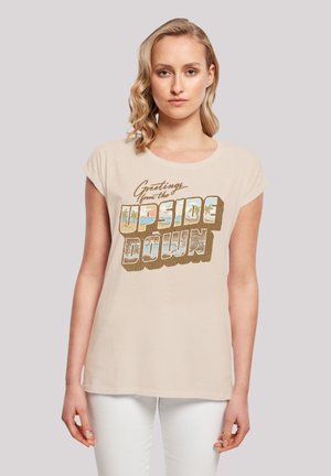 Helles beiges T-Shirt mit Rundhalsausschnitt und kurzen Ärmeln, das mit einem auffälligen grafischen Design verziert ist, auf dem "Grüße aus dem UPSIDE DOWN" steht.