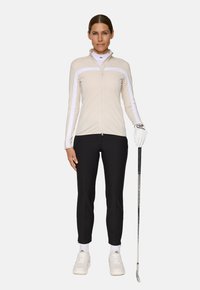 Maglione beige a maniche lunghe con zip e dettagli bianchi, abbinato a pantaloni aderenti neri e sneakers bianche. Un bastone da golf tenuto in una mano.