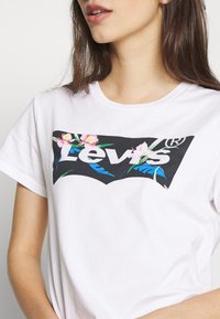 Valkoinen puuvillapaita, jossa on musta Levi's-logo, jota ympäröivät värikkäät kukka- ja lehtikuvat. Lyhyet hihat, pyöreä kaula-aukko, rento malli.