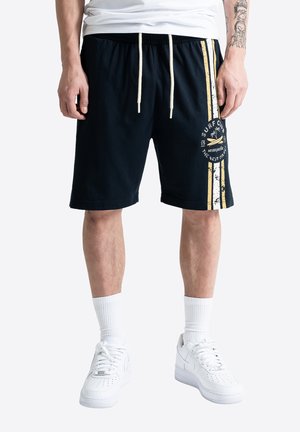 Homme portant un short noir à cordon avec des rayures jaunes et blanches et le logo du club de surf sur la cuisse, chaussettes blanches et baskets blanches.