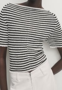 Närbild av en person som bär en kortärmad svart-vitrandig stickad topp och högmidjade vita byxor med dragkedja framåt, mot en enfärgad bakgrund.