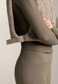 Beige textúrájú mellény műszőrme részletekkel, párosítva bordás, testhezálló olívazöld toppal és leggingszel. Oldalán pánt található, sima anyagfelülettel.