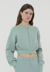 Felpa cropped zip-up verde menta con colletto rotondo, polsini a coste e una texture morbida, abbinata a pantaloni coordinati con coulisse.