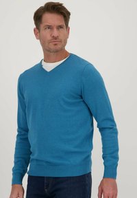 Blauwe v-hals trui van een lichte brei, met lange mouwen en ribgebreide manchetten en zoom. Geverfd over een wit t-shirt, tegen een neutrale achtergrond.