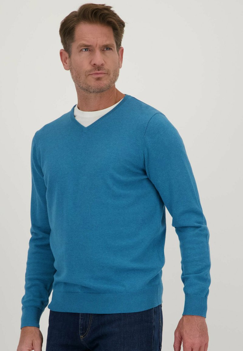 Blauwe v-hals trui van een lichte brei, met lange mouwen en ribgebreide manchetten en zoom. Geverfd over een wit t-shirt, tegen een neutrale achtergrond.