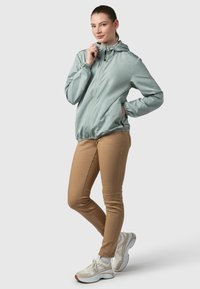 Gaiši piparmētru zaļa rāvējslēdzēja jaka ar kapuci, elastīgām aprocēm un sānu kabatām, kopā ar bēša krāsas šaurām biksēm un baltām sporta kurpēm.