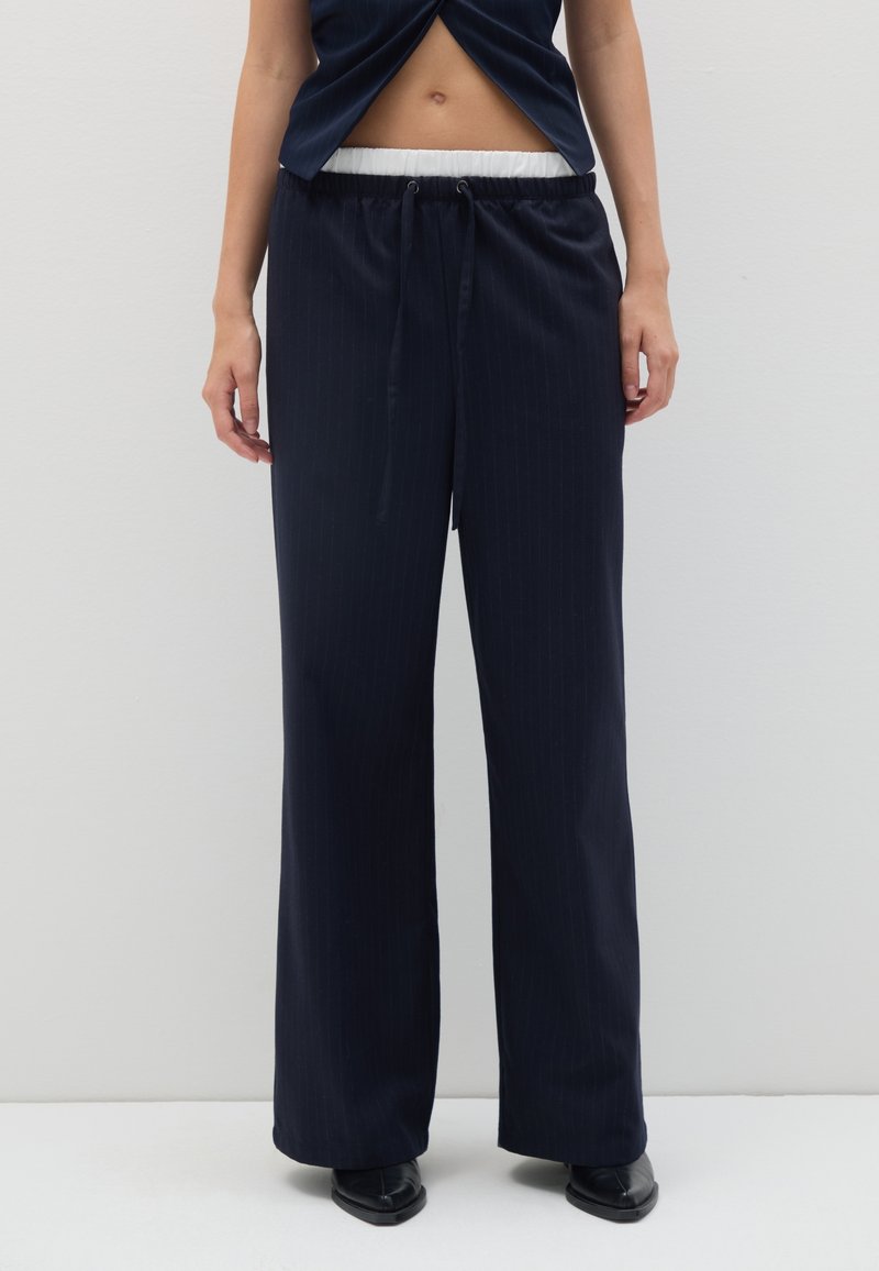 Femme portant un pantalon large à rayures noires avec une ceinture élastique blanche, assorti à des chaussures noires à bout pointu devant un mur uni.