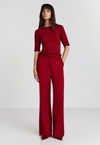 Frau, die vor einem schlichten Hintergrund steht und einen roten, kabelgestrickten Pullover mit kurzen Ärmeln, weite rote Hosen und spitze Schuhe trägt.