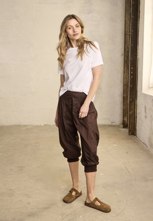 Mujer con cabello largo y rubio de pie en interiores, vistiendo una camisa blanca de manga corta, pantalones marrones sueltos y recortados, y zapatos marrones sin cordones.