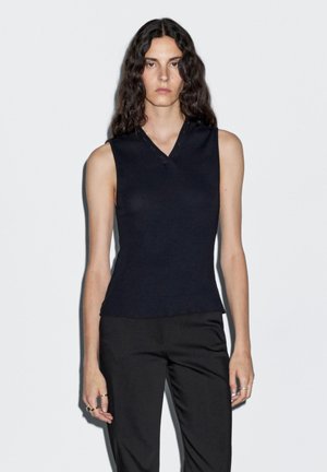 Navy sleeveless top with a ribbed texture, V-neck design, and button details at the neckline, paired with black trousers.