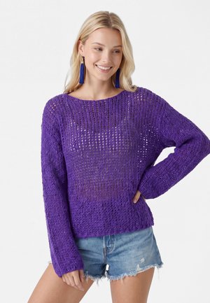 Kvinde med langt blond hår iført blå frynseøreringe, en løstsiddende lilla striktrøje og slidte denimshorts smilende med den ene hånd på hoften.