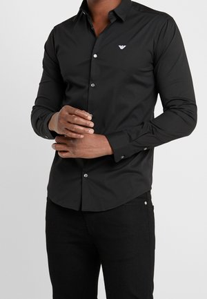 Mężczyzna ubrany w dopasowaną czarną koszulę na guziki z długim rękawem, z małym białym logo orła na lewej piersi, złożonymi rękami.