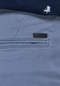 Gros plan sur la poche d’un pantalon bleu avec une étiquette de marque en cuir Jack & Jones et une chemise bleu marine avec un logo blanc brodé au-dessus.