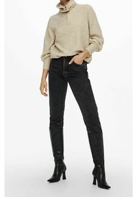 Beige gebreide pullover met een kraag met halve rits, zwarte high-waisted jeans en zwarte kniehoge haklaarzen. Gelaagd materiaal en aansluitend ontwerp.