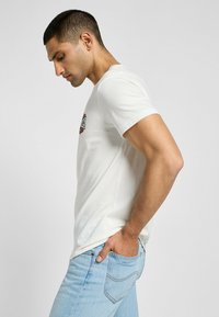 Biała koszulka z okrągłym dekoltem z grafiką na piersi, w połączeniu z jasnoniebieskimi dżinsami. Materiał wydaje się gładki i dopasowany.